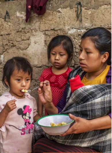 Mother in Guatemala feeds her daughter mashed bananas as another child looks on