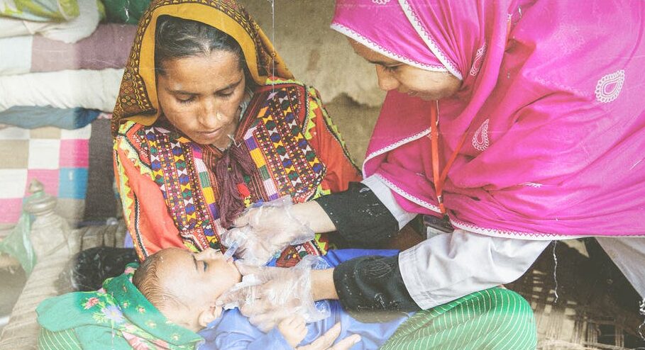 A healthcare worker in a pink headscarf and gloves administers oral vaccines to a baby lying on a woman's lap. The woman, dressed in colorful traditional clothing, watches attentively, highlighting the importance of child health in this modest indoor setting.
