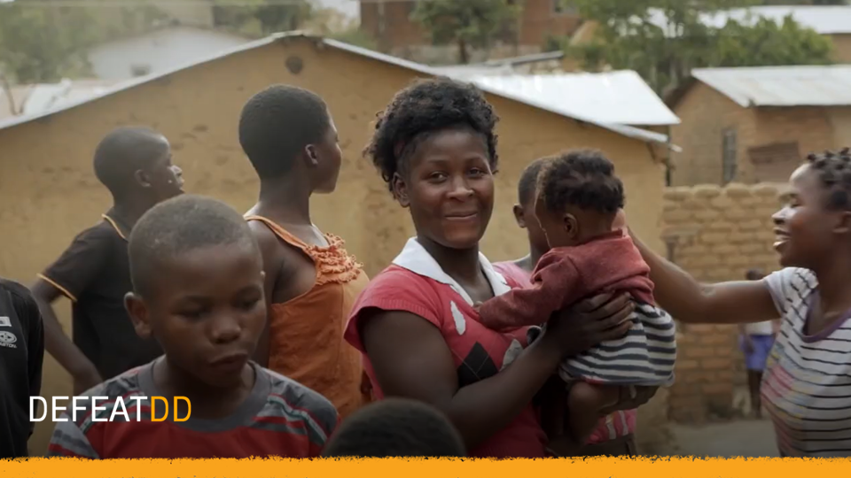 A smiling woman holds a baby in a community setting. Several people, including children, stand around her, engaging with one another. Brick buildings and a tree are in the background. The image has the text "DEFEATDD" at the bottom.
