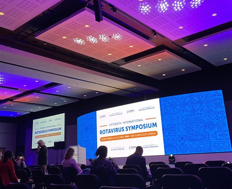 A conference room with people seated and a presenter on stage. Large screens display “Fifteenth International Rotavirus Symposium, 29 November–1 December 2023, Cape Town, South Africa” with event logos and blue decorative graphics.